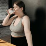 A-Woman-in-Gray-Tank-Top-Drinking-with-Her-Eyes-Closed-while-Standing-Near-the-Wooden-Table